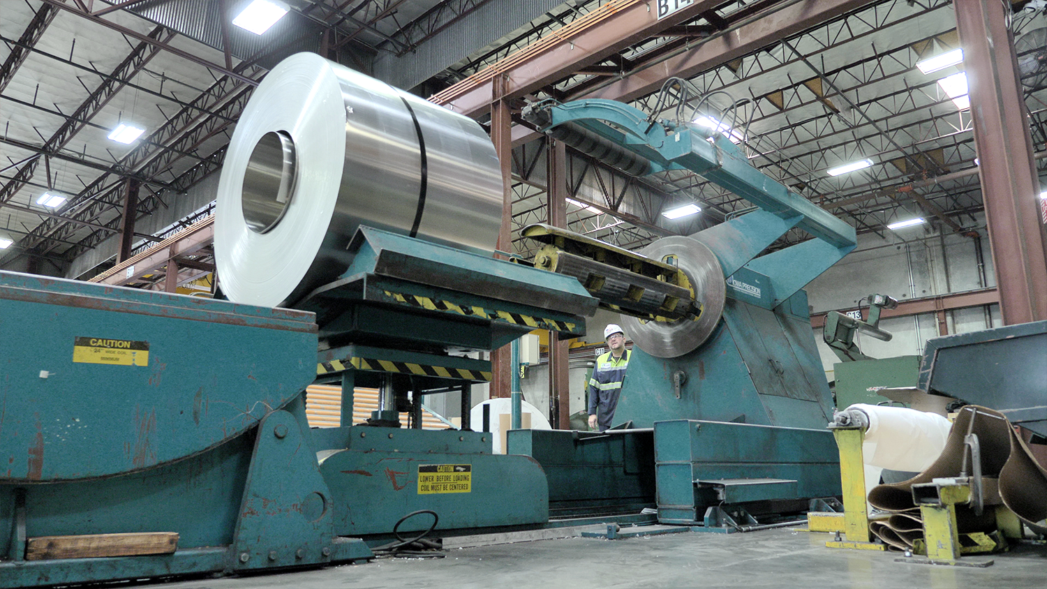 A steel coil is mechanically moved to a spindle while person observes.